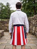 A man viewed from behind wearing a white dress shirt and a pleated kilt with vertical red and white stripes and a navy blue waistband, standing outdoors on a stone path with trees and stone walls in the background.