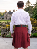 A man viewed from behind wearing a white long-sleeve dress shirt and a dark red pleated kilt with a black belt, standing outdoors in front of a stone building and garden.
