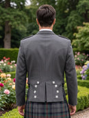 A man viewed from behind wearing a dark gray kilted jacket and a tartan kilt in a garden setting.