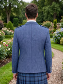 A man viewed from behind wearing a textured blue herringbone jacket and a blue tartan kilt, standing on a gravel path in a lush green garden.